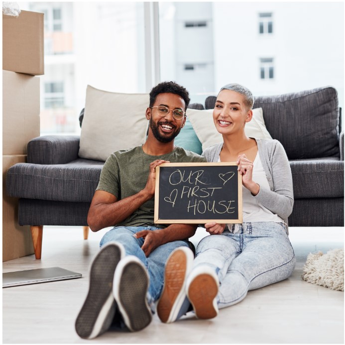 A smiling couple sits on the floor of their new home, holding a chalkboard sign that reads "Our First House" with hearts drawn on it. Behind them are a gray couch and unpacked moving boxes, indicating they have just moved in.