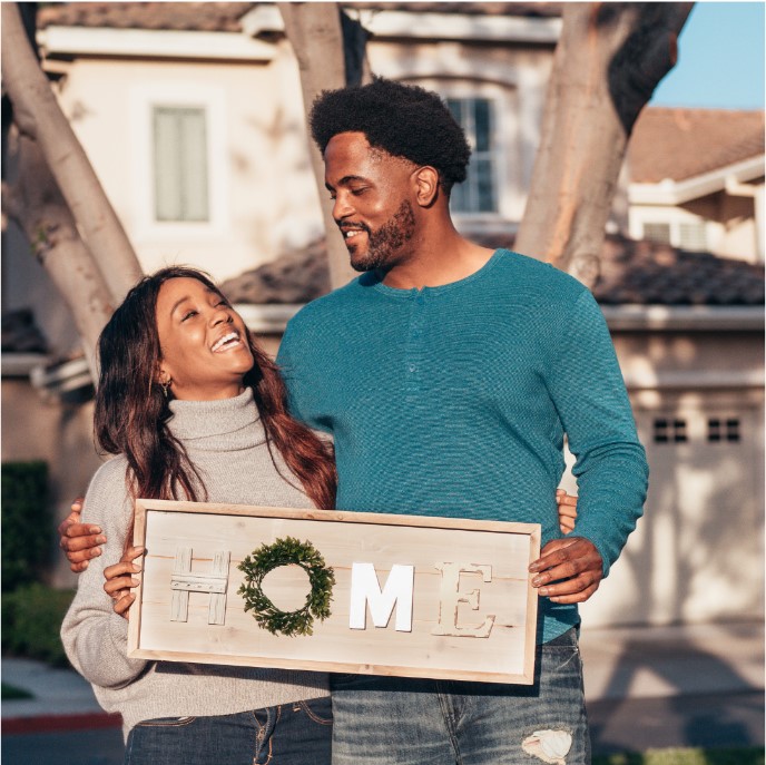 A smiling couple stands outside a house holding a sign that says "HOME.