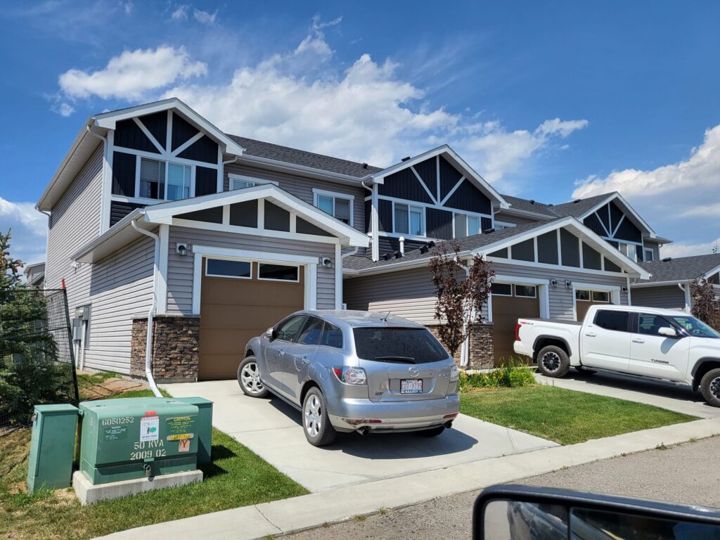 2 Townhouses along a street with 2 cars in their garages on a sunny day with well manicured green grass between the 2 houses.