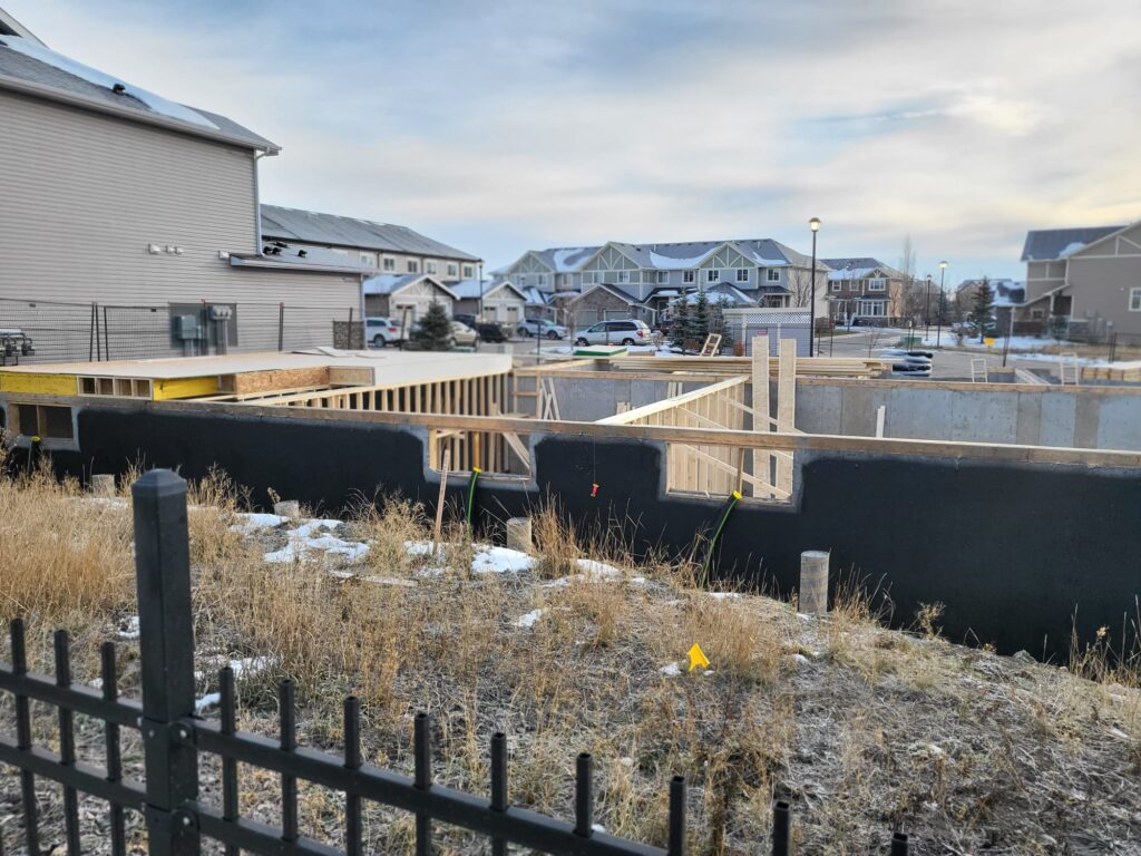 A house is being constructed with short wood walls coming from the foundational base of the house. The construction is surrounded by townhomes with cars infront of them.