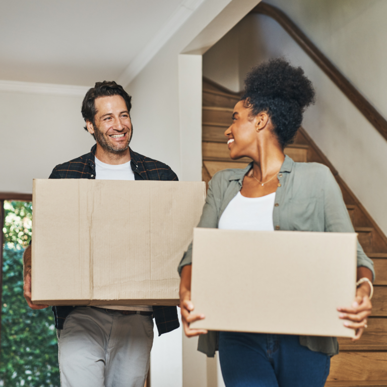 A man and woman smile at each other as they carry large cardboard boxes, clearly in the midst of moving house. They appear to be in a home with a wooden staircase in the background. The man is wearing a plaid shirt, while the woman sports a green shirt and white top.