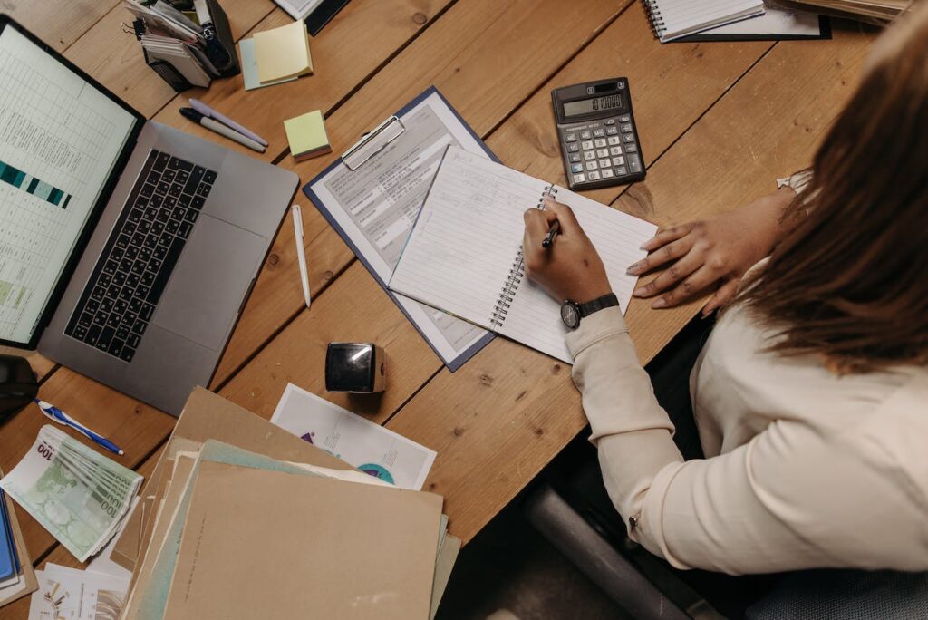 A person sits at a wooden desk, writing in a notebook. The desk is cluttered with various items including a laptop displaying charts, a mortgage calculator, papers, sticky notes, and euro banknotes. The individual wears a watch and a light-colored shirt.