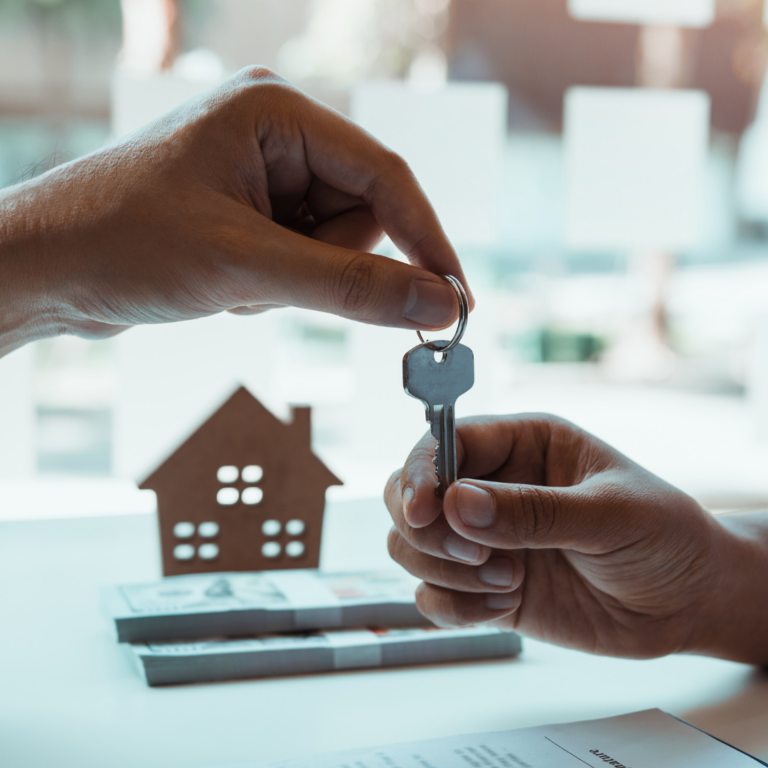 Two hands exchanging a silver key indoors, with one hand giving and the other receiving. In the background, there is a small wooden house model and some documents on a white table.