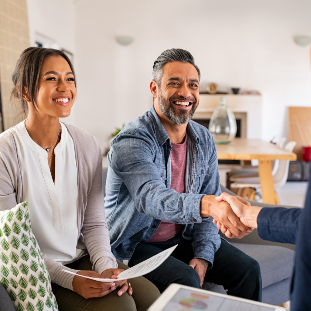 A smiling couple is indoors, sitting on a couch. The man, wearing a denim shirt, is reaching out to shake hands with someone off-frame. The woman, in a white blouse and cardigan, holds a document. The setting appears to be a friendly meeting or consultation in a cozy living room.