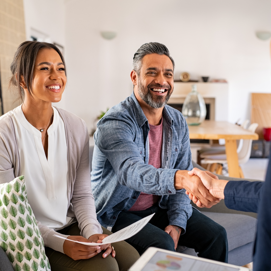 A smiling couple is indoors, sitting on a couch. The man, wearing a denim shirt, is reaching out to shake hands with someone off-frame. The woman, in a white blouse and cardigan, holds a document. The setting appears to be a friendly meeting or consultation in a cozy living room.