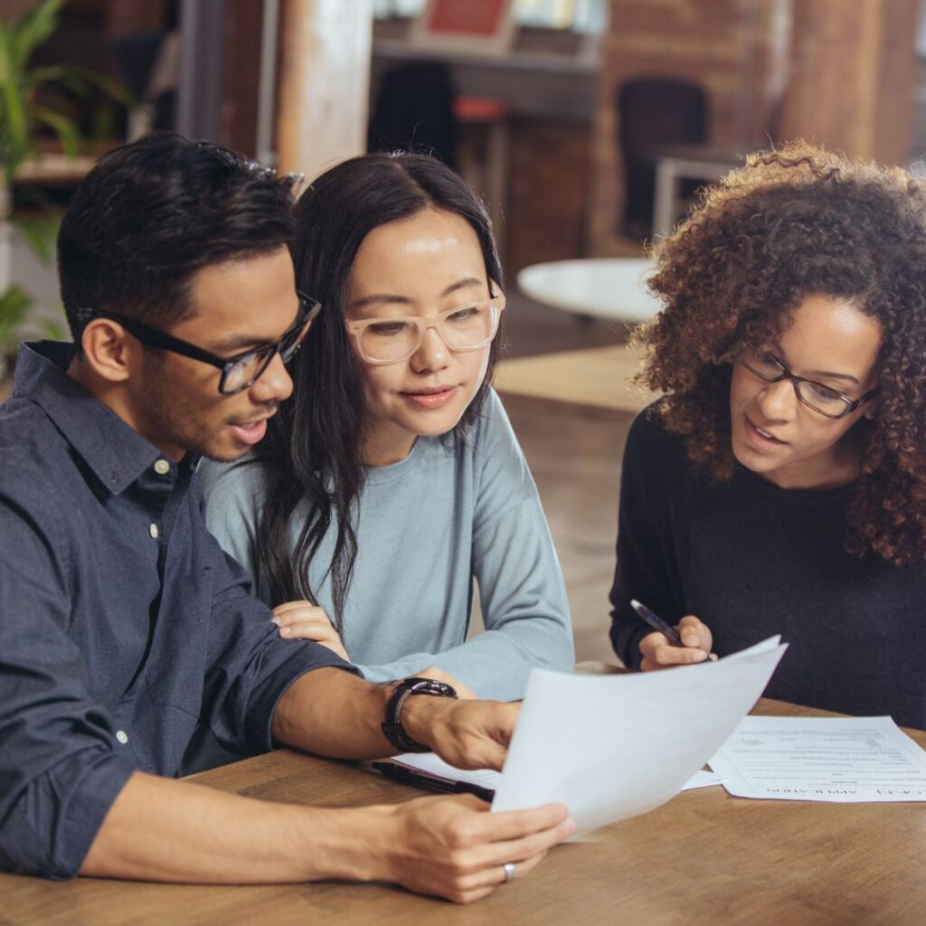 Three people are sitting at a wooden table in a casual office setting, focused on documents and papers in front of them. They are wearing glasses and are engaged in a collaborative discussion or review session about credit score approval to buy a home.