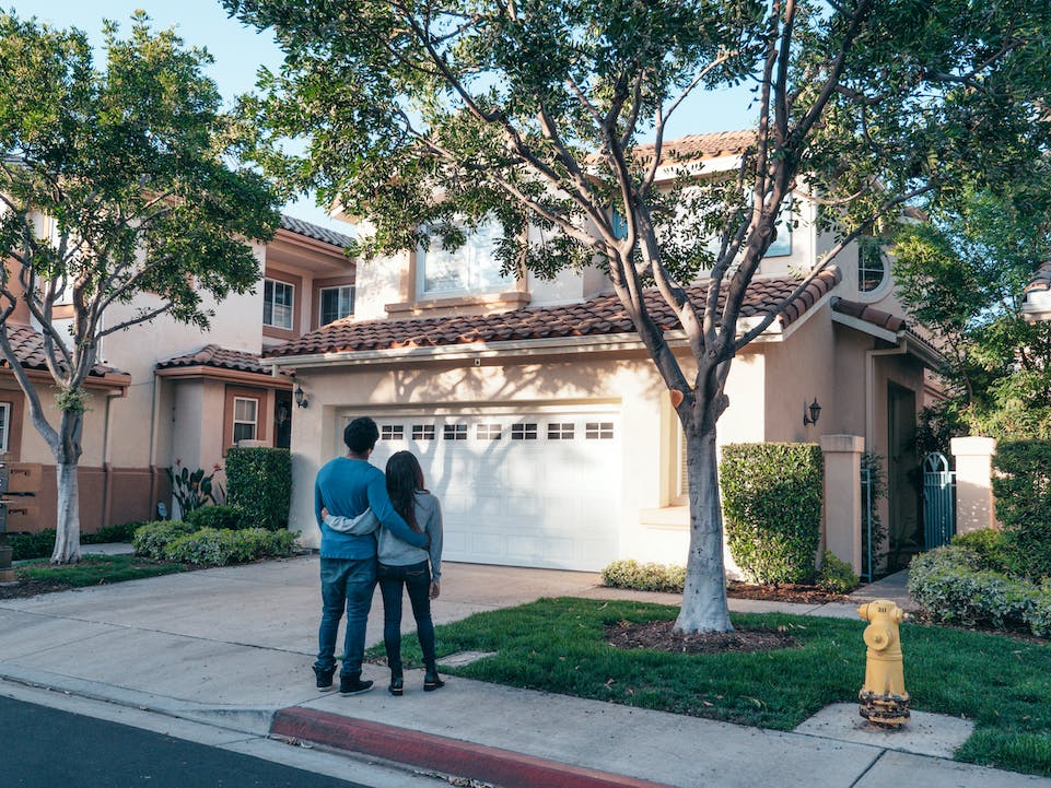 A couple stands on the sidewalk with their backs to the camera, looking at a two-story house with a well-maintained yard and trees. They have their arms around each other. A yellow fire hydrant is visible on the grass near the curb.