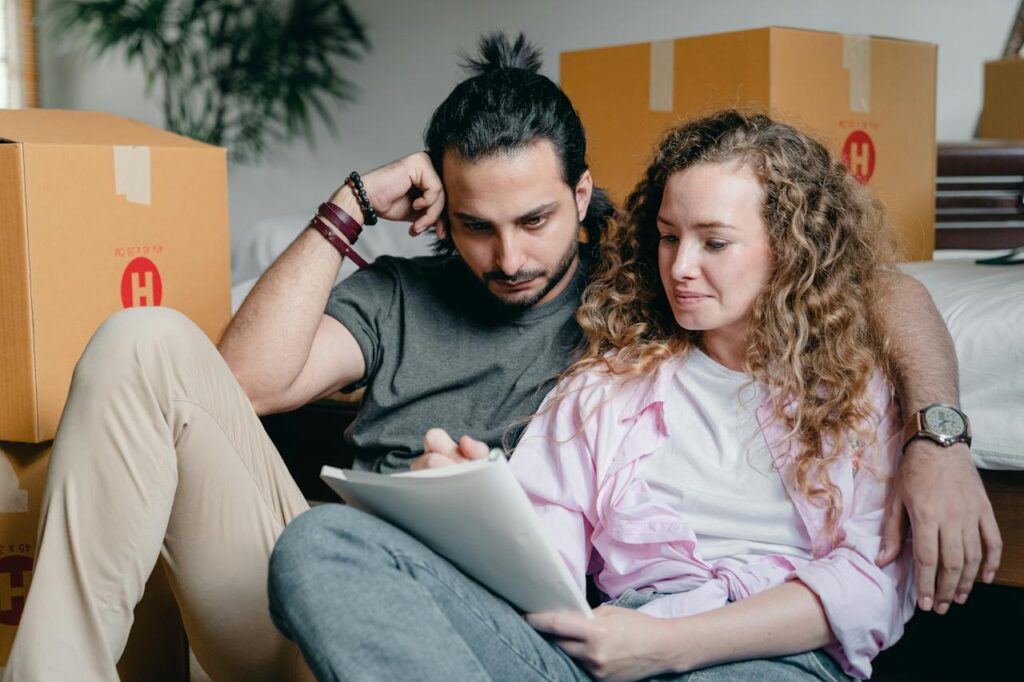 A couple sits on the floor among moving boxes. The man, with long hair tied back, leans against the woman, who has curly hair. They are both looking at a piece of paper that the woman is holding, making the descision of whether to Buy or To Rent?