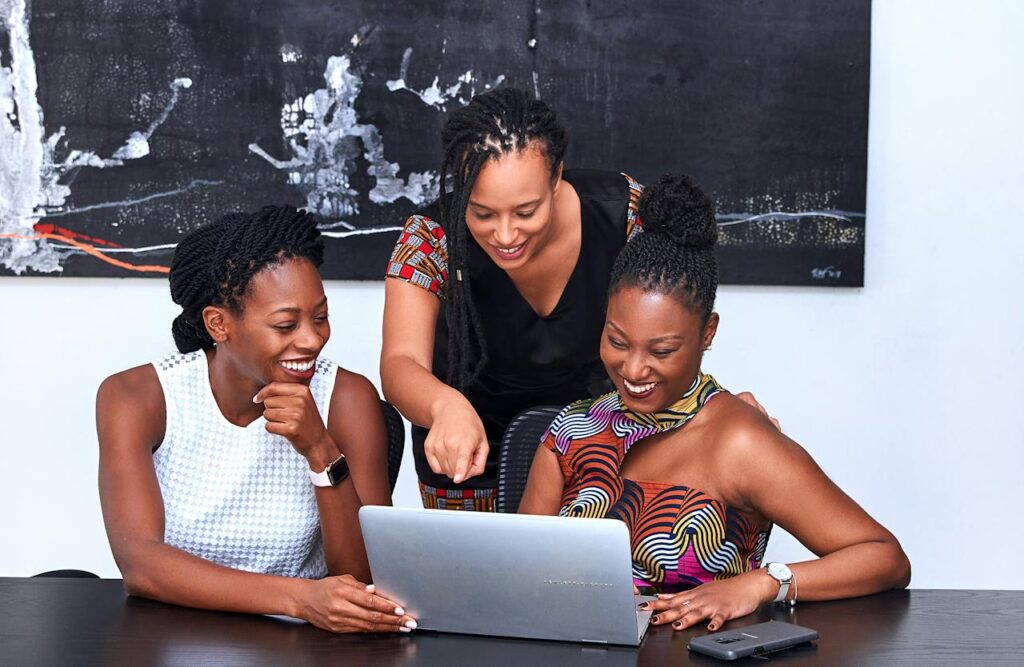 Three women are gathered around a laptop, looking at the screen and smiling. One woman, standing in the middle, is pointing at the screen. They appear to be in an office setting with a large abstract painting on the wall behind them.