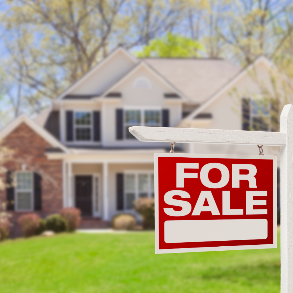 A "For Sale" sign hangs in front of a two-story house with a mix of brick and siding exterior, black shutters, and an inviting front porch. The well-maintained lawn and trees in the background suggest it is a suburban home on a sunny day.