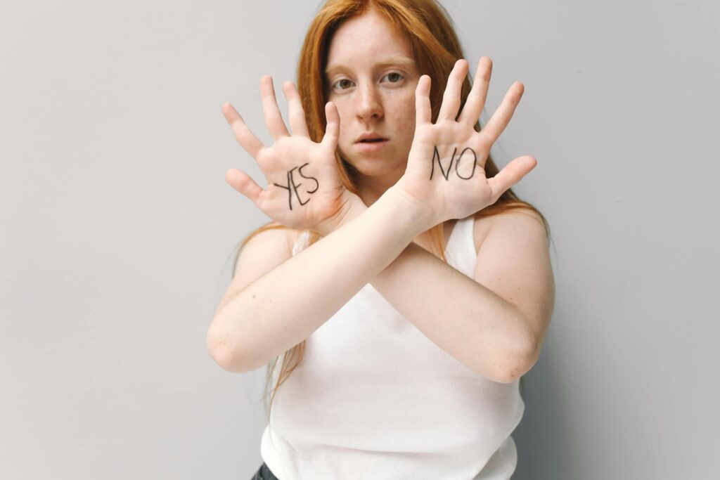 A person with long red hair and wearing a white tank top holds up their hands with crossed arms. One hand has "YES" written on the palm, and the other has "NO," set against a plain gray background.