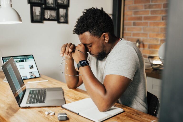 A person sits at a wooden desk with their eyes closed and hands clasped, resting their forehead on their hands. On the desk, there is an open notebook, a laptop, a smartphone, earbuds, and glasses. The background includes a brick wall and framed pictures.