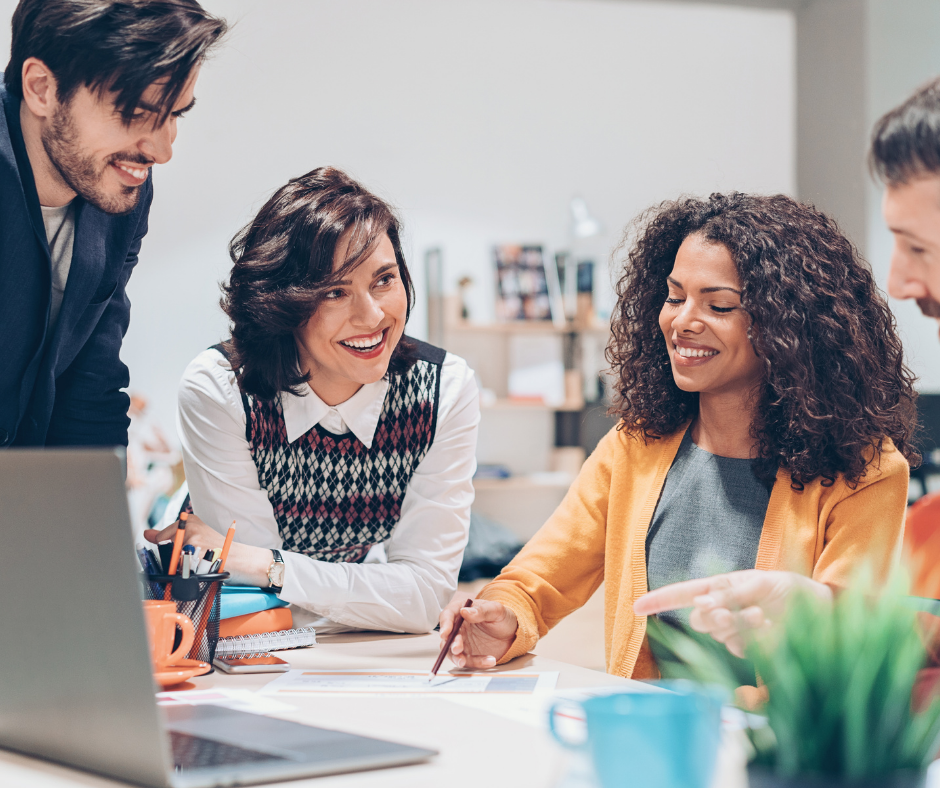 A group of four colleagues sit around a table, engaged in a discussion. They are smiling and appear to be planning or brainstorming. One person points at a paper on the table.