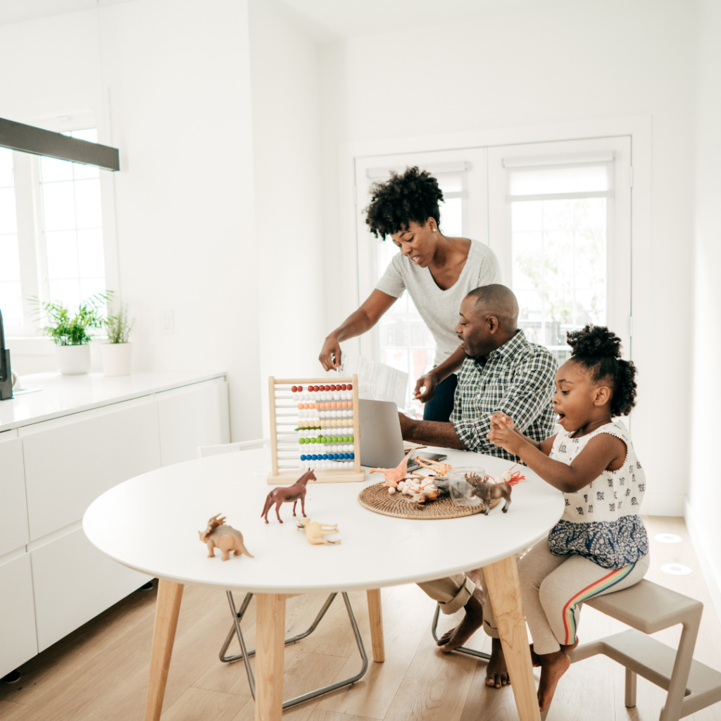 A family of three is playing at a white table in a bright, modern kitchen. The child is using an abacus, while the parents watch and engage with the child. Toy dinosaurs are scattered on the table. The scene is filled with natural light from nearby windows.