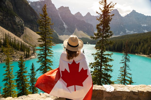 A lady sitting by a scenic lake and mountain scene is wrapped in a Canadian flag.