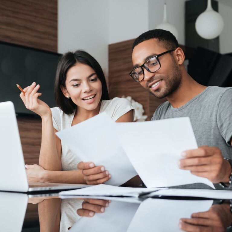 A man and woman looking at 2 documents while sitting by a computer.