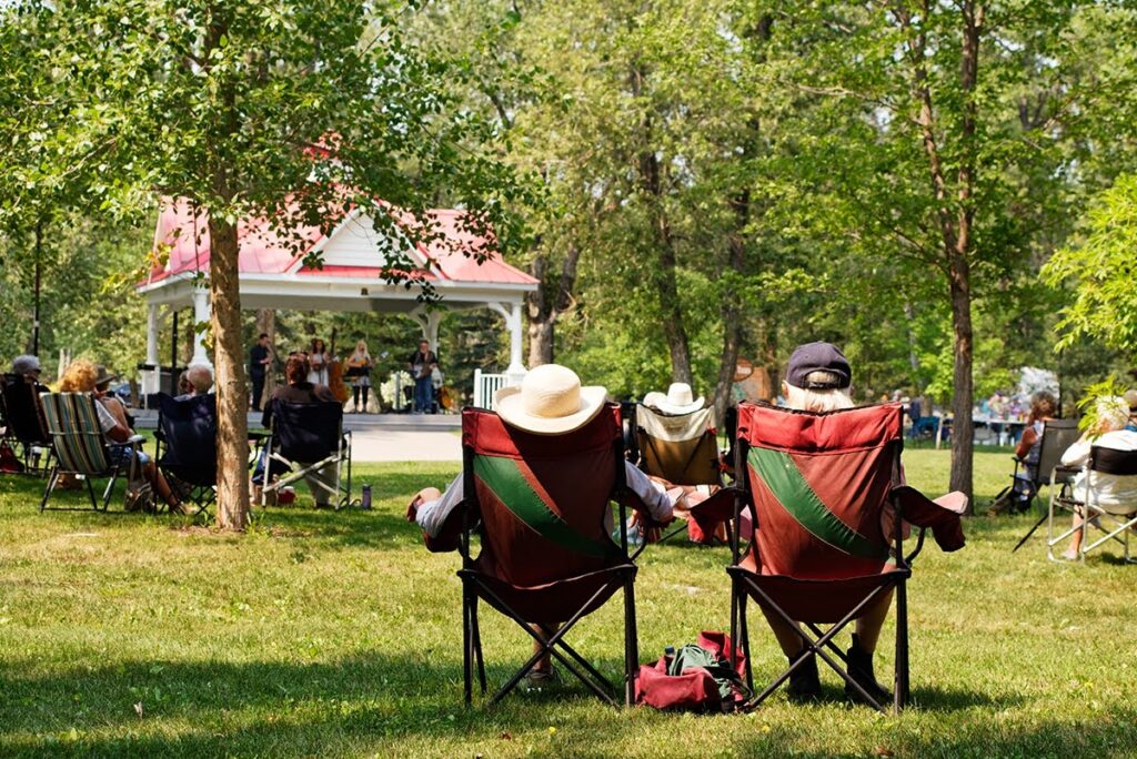 A man and woman sitting on lawn chairs in a park on a sunny day, watching a muci band playing on stage, surrounded by other people.