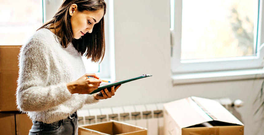 A woman stands in a room filled with packing boxes, writing on a clipboard with a house moving checklist. She is wearing a fuzzy white sweater and jeans. Sunlight streams in through a window behind her, creating a warm atmosphere.